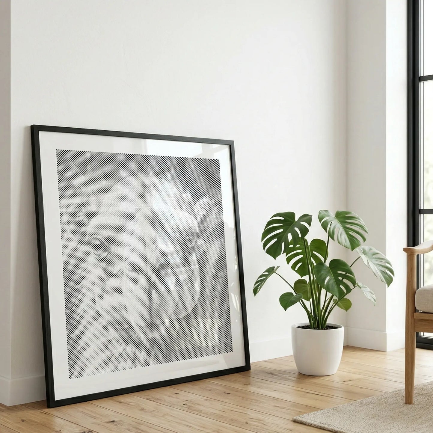 A large black-framed black and white art print of a camel's face leaning against a white wall on a light wood floor, next to a potted green plant and a wooden chair. Camel | Dot Painting Kit