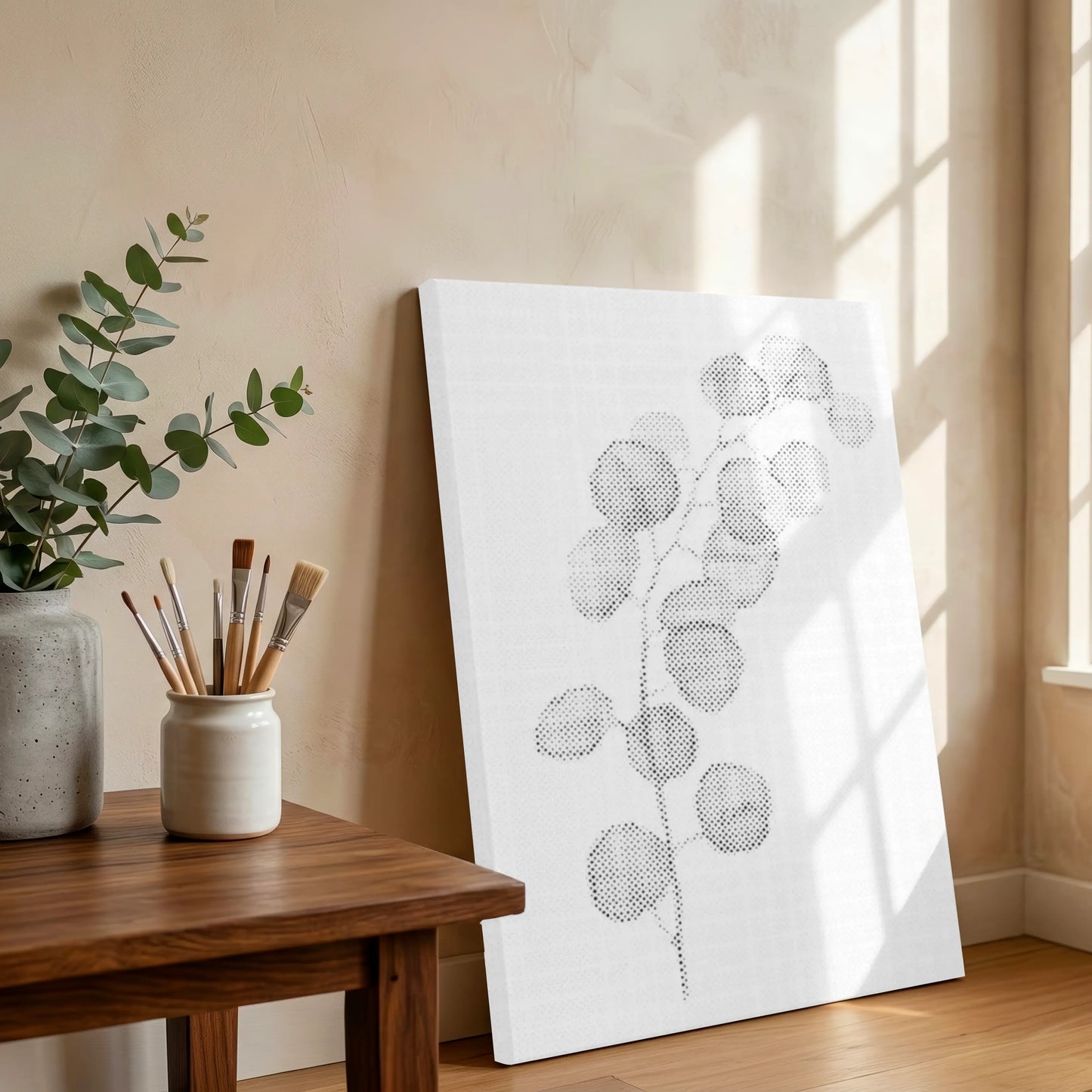White canvas with black floral design on a wooden table with a plant and paintbrushes. Eucalyptus Branch | Dot Painting Kit
