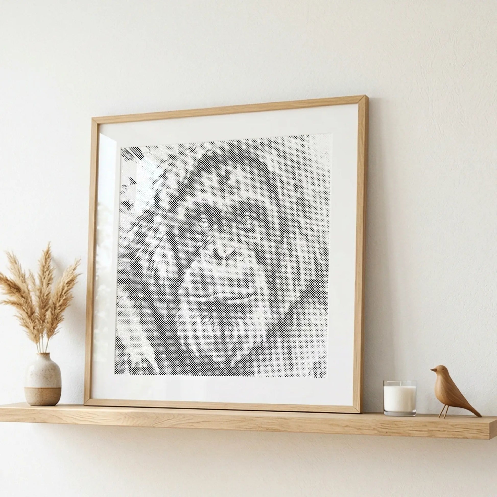 A framed black and white portrait print of an orangutan hanging on a light wall above a wooden shelf with decorative dried grass, a candle, and a small bird figurine. Orangutan | Dot Painting Kit